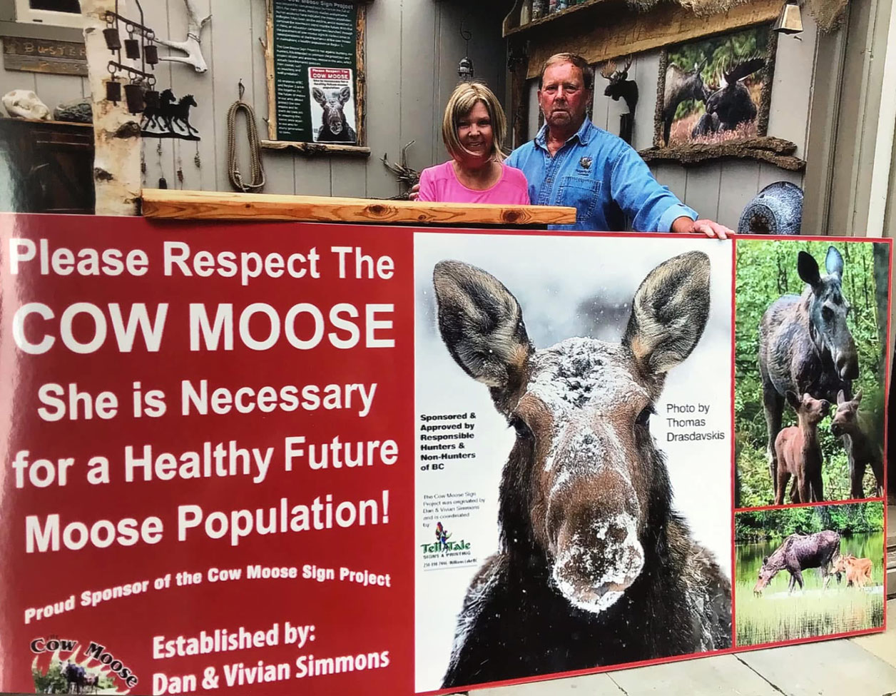 Dan and Vivian Simmons, founders of the Cow Moose Sign Project in British Columbia stand behind one of their signs
